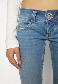 Jasnoniebieskie dżinsy o dopasowanym kroju, wysoki stan i zapięcie na dwa guziki. Posiadają styl pięciu kieszeni z subtelnym przeszyciem.