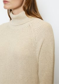 Femme portant un pull beige en maille à col roulé avec manches longues, montrant les détails de l'épaule et du cou sur un fond blanc.
