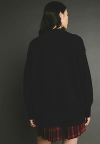Pulover negru supradimensionat din lână tricotată, cu manșete și tiv texturat. Purtați peste o fustă plisată cu carouri roșii. Design simplu și texturat, cu umeri căzuți.