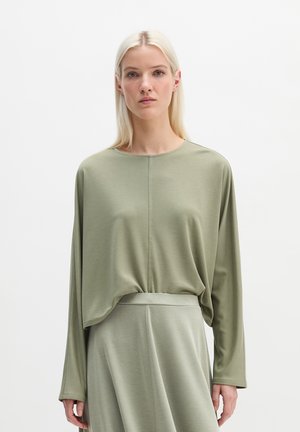 Vrouw met lang blond haar, gekleed in een losse lichtgroene top met lange mouwen die in een bijpassende hooggetailleerde rok is gestopt, staand tegen een witte achtergrond.