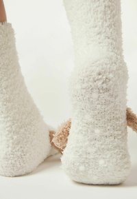 Calzini bianchi morbidi con una superficie testurizzata, dotati di accenti in peluche beige che richiamano le orecchie di un animale. Dotati di punti antiscivolo sulle suole per una maggiore aderenza.
