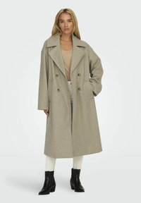 Lichtgrijze dubbelzijdige wollen jas met grote revers, twee voorkruiken en oversized pasvorm, gedragen over een beige top en witte broek.