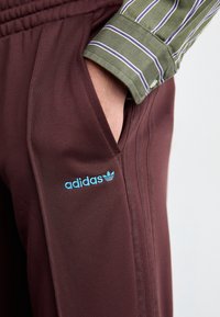 Maroon athletic pants with side pockets, featuring blue embroidered Adidas logo and textured fabric. Striped shirt visible at the cuff.
