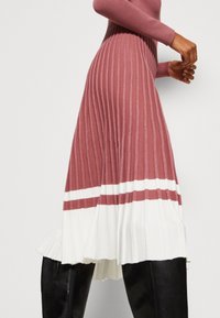 Ribbed midi-mekko vaaleanpunaisena, jossa on valkoiset raidat helmassa, saalen alareuna ja pehmeä tekstuuri. Päällä mustat nahkahousut.