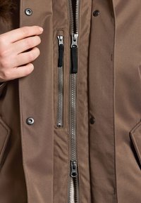 Bruine buitenkleding met een gladde textuur, voorzien van meerdere ritsen en drukknoopjes. Close-up van een hand op de stof, die de ontwerpdetaillering laat zien.