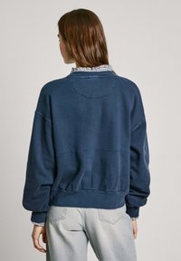 Marinblå oversize sweatshirt med ribbade mudd och nederkant, med detaljer i form av en cirkulär ok och mjukt, texturerat tyg. Bärs över en skjorta med krage.