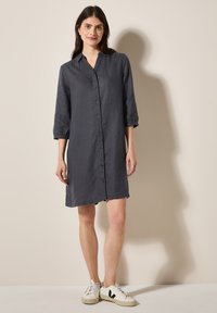 Tumma harmaa pellavasta valmistettu shirt dress, jossa on kaulus, nappikiinnitys ja kolme neljäsosaa hihoista; yhdistetty valkoisiin tennareihin ja neutraalin taustan kanssa.