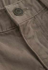 Lichtbruin materiaal, close-up van een broek met een metalen knoop, stiksel details en een gestructureerde oppervlakte.