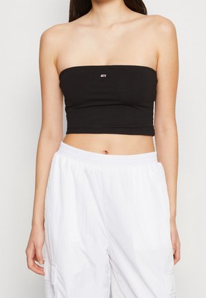 Vrouw met een zwarte strapless cropped top en losse witte broek, van schouders tot heupen afgebeeld tegen een effen achtergrond.