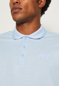 Polo de color azul claro hecho de tejido texturizado con cuello, tapeta de tres botones y logo bordado "AM" en el pecho.