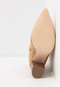 Chaussure à talon haut en suede beige avec une pointe en amande et une semelle en caoutchouc brun texturée, présentant un petit talon empilé.