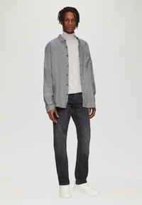 Grijze button-up shirt over een lichte turtleneck, gecombineerd met donkere jeans en witte sneakers. Kenmerken zijn onder andere een borstzak en een relaxte pasvorm.