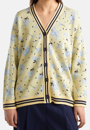 Cardigan jaune à motif floral bleu, bordure rayée marine et boutons noirs, porté avec un pantalon marine.