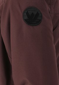 Bordeauxkleurige stof met een gestructureerd oppervlak en een ronde zwarte patch met een gestileerd "W" logo, gelabeld "WHISTLER DENMARK."