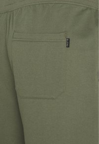 Joggers en tissu vert olive avec une poche arrière unique en forme arrondie et des détails cousus. Petite étiquette 'BLEND' sur la poche.