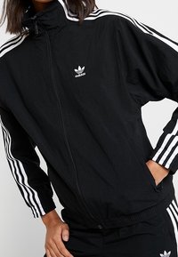 Černá sportovní bunda Adidas s vysokým límcem, předním zipem a třemi bílými pruhy podél rukávů. Lehký materiál s elastickými manžetami.