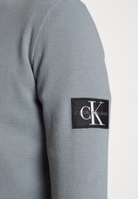Graues Langarmshirt mit strukturierter Oberfläche, versehen mit einem schwarzen rechteckigen Patch mit dem Logo "Calvin Klein Jeans" und weißer Schrift.