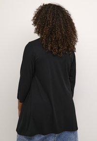 Haut noir à manches trois-quarts avec une silhouette fluide, fabriqué en matériau doux. Présente un ourlet arrondi, porté sur un denim bleu.