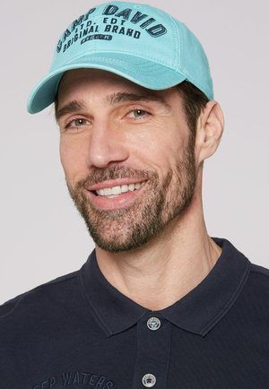 Turquoise baseball cap with embroidered text, worn by a person in a dark navy polo shirt. Visible stitching and a button detail on the collar.