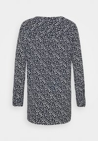 Blouse noire à manches longues avec des pois blancs irréguliers, coupe ample et ourlet arrondi, vue de dos.