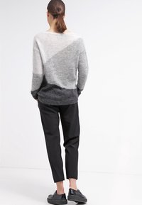 Donna con ponytail castano che indossa un dolcevita grigio sfumato, pantaloni cropped neri e sneakers con plateau nere, in piedi di fronte a uno sfondo bianco.
