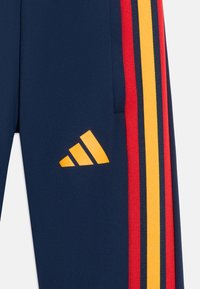 Námořnické atletické kalhoty s červenými a žlutými pruhy po stranách, s žlutým logem Adidas na levé straně a hladkou, lesklou texturou látky.