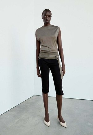Donna con top smanicato color taupe, pantaloni neri al ginocchio e tacchi bianchi a punta, in piedi davanti a un muro bianco semplice e pavimento grigio.