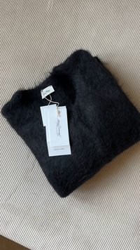 Črn pleten kardigan iz puhastega materiala z gumbi in priloženimi etiketi za nego. Zložen na teksturirani svetli podlagi.