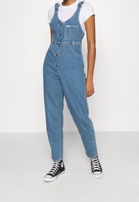 Denimöveraller med en ljusblå tvätt, justerbara axelremmar, framknappar, sidofickor och design med avsmalnande ben. Bärs med svarta sneakers.