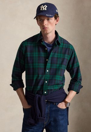 Jeune homme portant une chemise à carreaux vert foncé et bleu marine, une casquette bleu marine, un jean et une montre, avec un pull bleu marine noué autour de la taille.