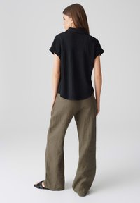 Camicia nera a maniche corte con un motivo testurizzato, abbinata a pantaloni in lino verde oliva dal taglio ampio e sandali neri.