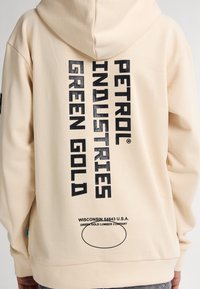 Sudadera beige con capucha ajustable. Presenta un texto en negrita de color negro: "PETROL INDUSTRIES GREEN GOLD" y una dirección de Wisconsin en la parte posterior.