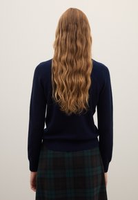 Marineblauer Strickpullover mit langen Ärmeln und einem gerippten Bund. Kombiniert mit einem dunkelgrünen und schwarzen karierten Rock.