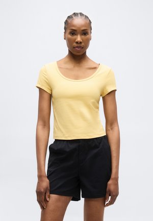 Νεαρή γυναίκα με πλεγμένα μαλλιά, φορώντας στενό κίτρινο κοντομάνικο μπλουζάκι και φαρδιά μαύρα σορτς, στέκεται μπροστά σε απλό ανοιχτό φόντο.