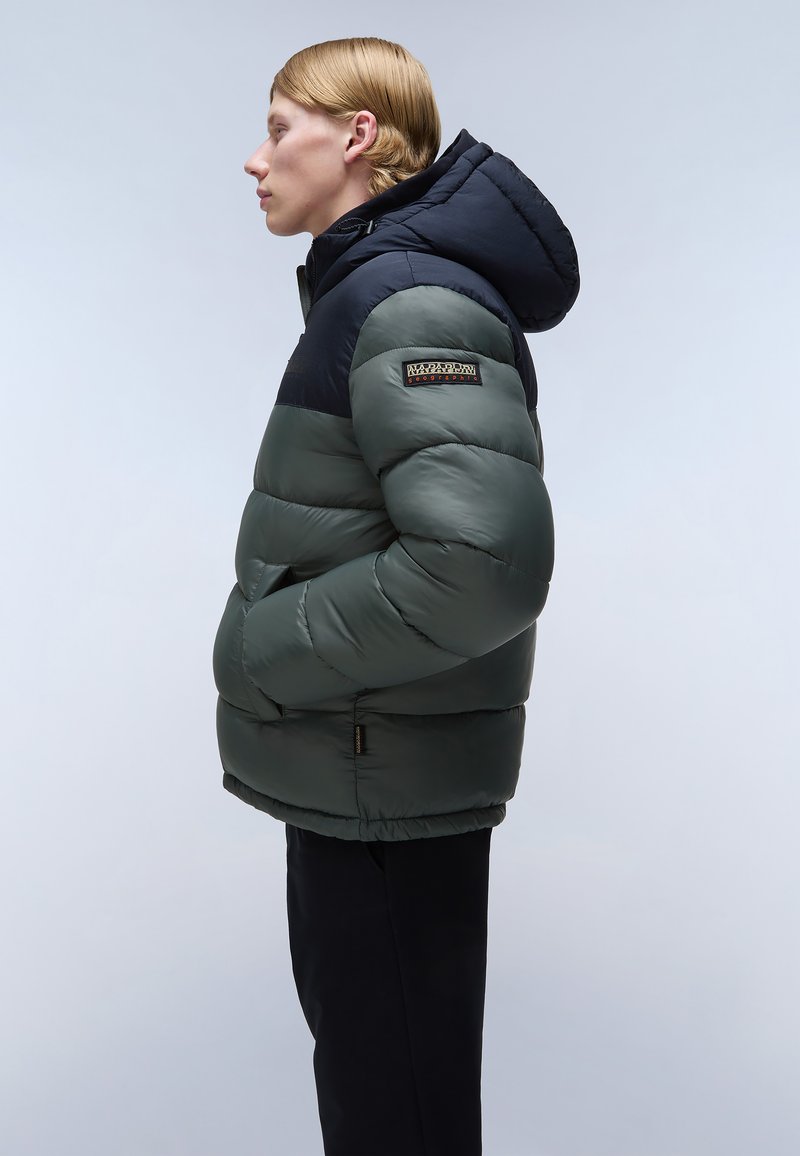 Puffermantel in Grün und Marineblau, mit Kapuze, gestepptem Design, Reißverschlusstaschen und einem Logo-Patch auf der linken Schulter.