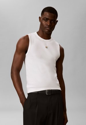 Mężczyzna ubrany w białą, bezrękawną żebrowaną koszulkę z małym wielokolorowym logo, czarne spodnie i czarny pasek, stoi na szarym tle.