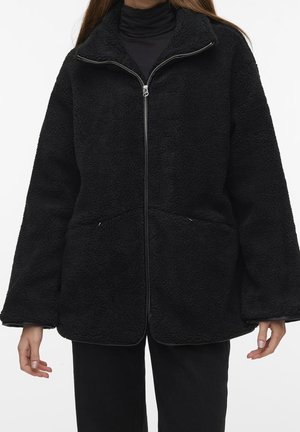 Kobieta ma na sobie czarną polarową kurtkę zapinaną na zamek z wysokim kołnierzem, na czarnym golfie i czarnych spodniach, stoi na białym tle.