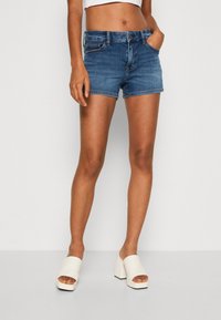 Žena v modrých džínech šortkách, bílém zkráceném topu a bílých sandálech na podpatku stojící na jednobarevném pozadí.