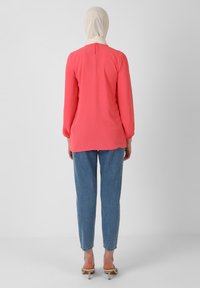 Blusa corallo con maniche lunghe e polsini elasticizzati, con chiusura a bottoni dietro, abbinata a jeans alta vita azzurri e tacchi beige.