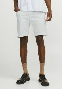 Pantalones cortos de algodón blanco con dobladillos enrollados, bolsillos laterales, combinados con zapatos negros y calcetas marrones. Diseño minimalista, textura suave.