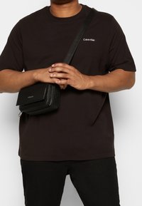 Svart bomullströja med en vit "Calvin Klein"-logotyp. En liten svart axelväska med ett strukturerat mönster hänger från axeln.