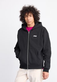 Black zippered hoodie made of cotton with a kangaroo pocket and white logo on the left chest. Worn over a pink shirt, styled with striped pants.