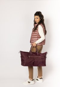 Bordowa torba podróżna z podwójnymi uchwytami, zamkiem błyskawicznym oraz wytłoczonym logo, wykonana z trwałego materiału. Model ubrany w jasną kamizelkę i spodnie.