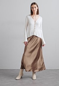 Λευκό ριμπ μακρυμάνικο τοπ με κουμπιά, σε συνδυασμό με καφέ σατέν μάξι φούστα. Το μοντέλο στέκεται σε μπεζ μπότες πάνω σε γκρι δάπεδο.