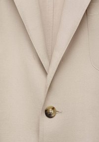 Beige linen blazer featuring a pointed lapel, single button closure with a marbled finish, and a chest pocket detail.