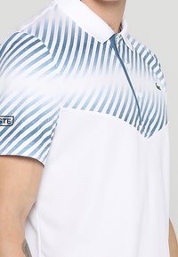 Vit kortärmad pikétröja med ett blårandigt mönster upptill och krage. Har en logotypetikett på ärmen. Mjuk textur.