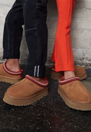 Scarpe slip-on in camoscio color fango con bordo decorato rosso e bianco. Suole di gomma spesse e forma arrotondata. Indossate con pantaloni neri e arancioni.