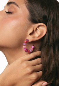Pendientes de aro de cuentas en tonos morado y rosa con acentos dorados, que presentan una textura variada y formas redondeadas, lucidos por una modelo que destaca una oreja.