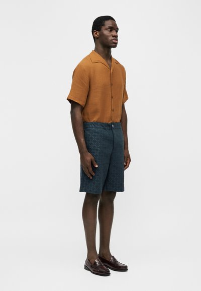 Άνδρας που στέκεται, φορώντας πουκάμισο βραχείας μανσέτας χρώματος σκουριά, σορτς με ναυτικό μπλε μοτίβο και σκούρα καφέ μοκασίνια, σε λευκό φόντο.