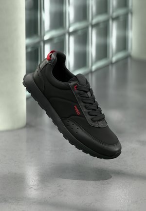 Musta urheilutennari punaisilla yksityiskohdilla ja Hugo-logolla, leijuu harmaan kiillotetun lattian yläpuolella lasitiiliseinän ja valkoisen pylvään lähellä.